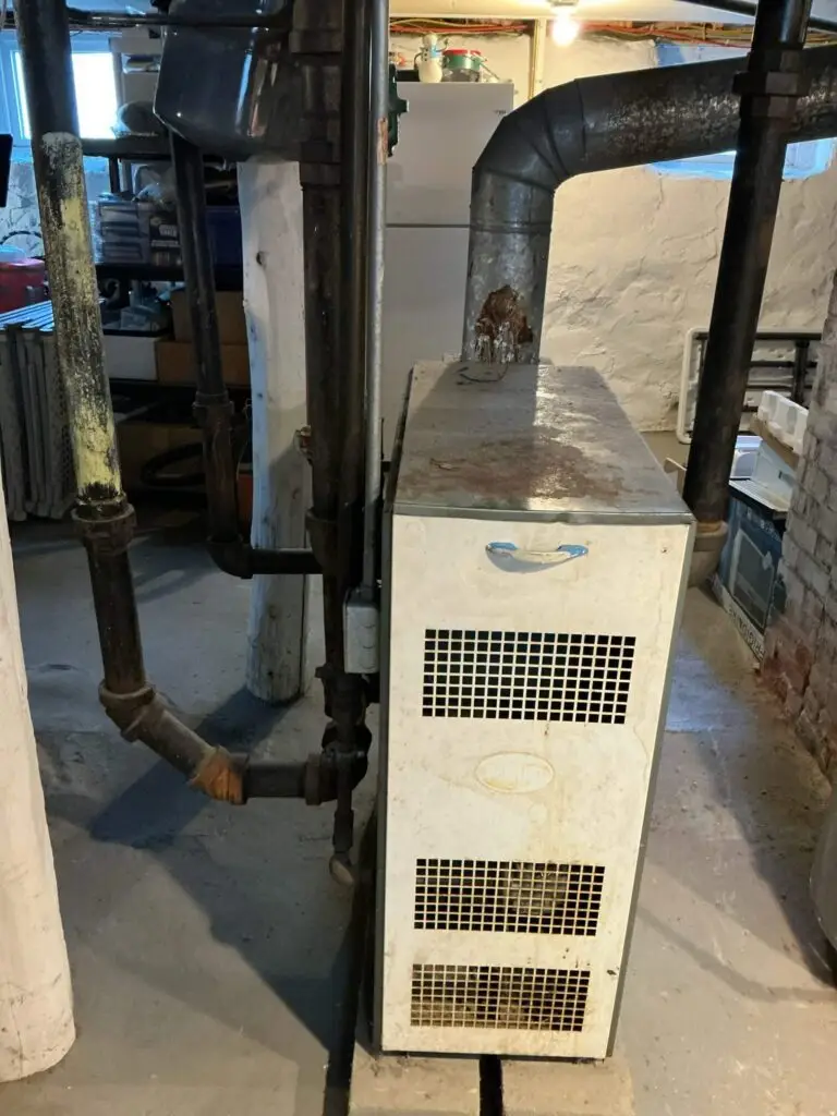 Older basement heating unit with exposed pipes and rusted metal ductwork before replacement